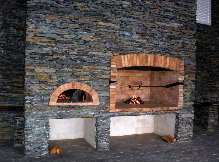 Kamenná stěna s pizza pecí a krbem pod cihlovými klenbami, v obou hoří oheň